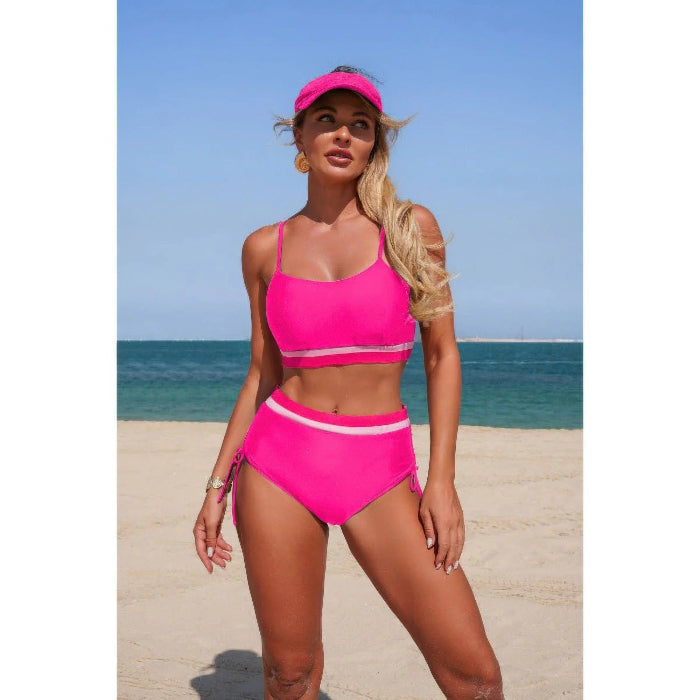 A woman modeling a bright pink two-piece bikini with a matching visor on a sandy beach with the ocean and blue sky in the background.