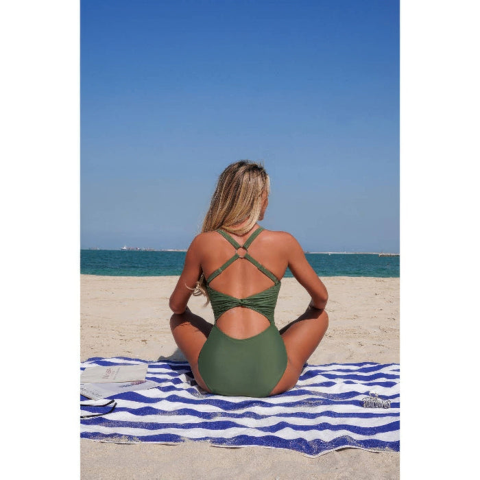 A woman sits on a blue and white striped beach towel on a sandy beach, viewed from behind, wearing an olive green one-piece swimsuit with crisscross back straps.