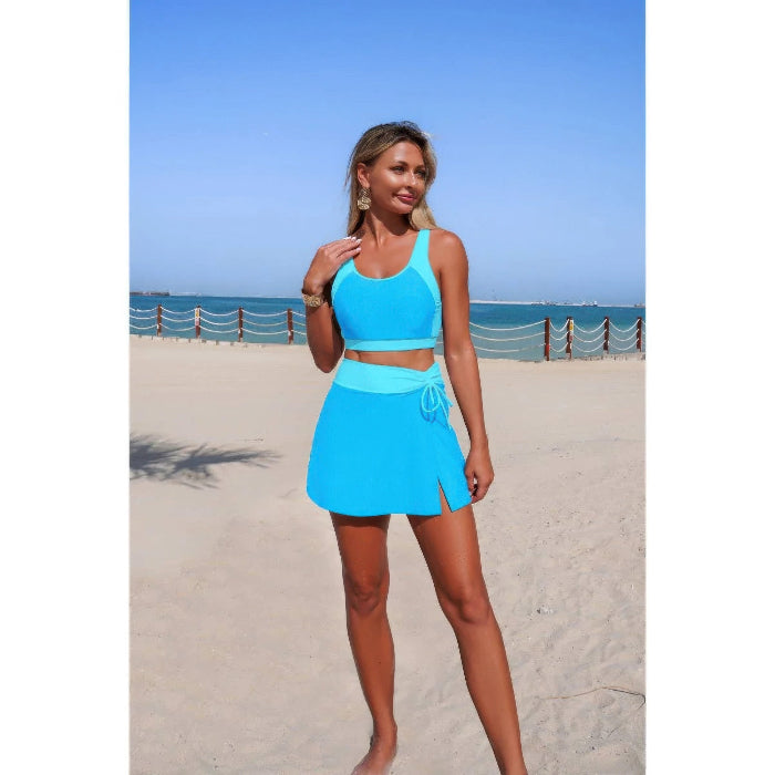 A woman modeling a bright turquoise blue two-piece swimsuit on a sandy beach with the ocean and blue sky in the background.
