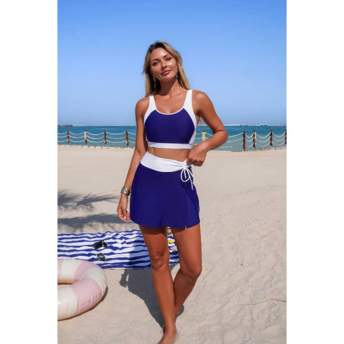 A woman modeling a two-piece swimsuit on a sandy beach. The set features a royal blue and white crop top with a white waist tie and matching high-waisted royal blue shorts.