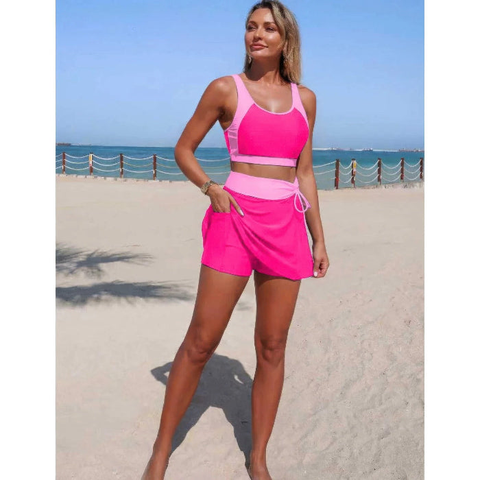 A woman modeling a two-piece athletic set consisting of a sports bra and matching shorts in bright pink and light pink, standing on a sandy beach with the ocean and blue sky in the background.
