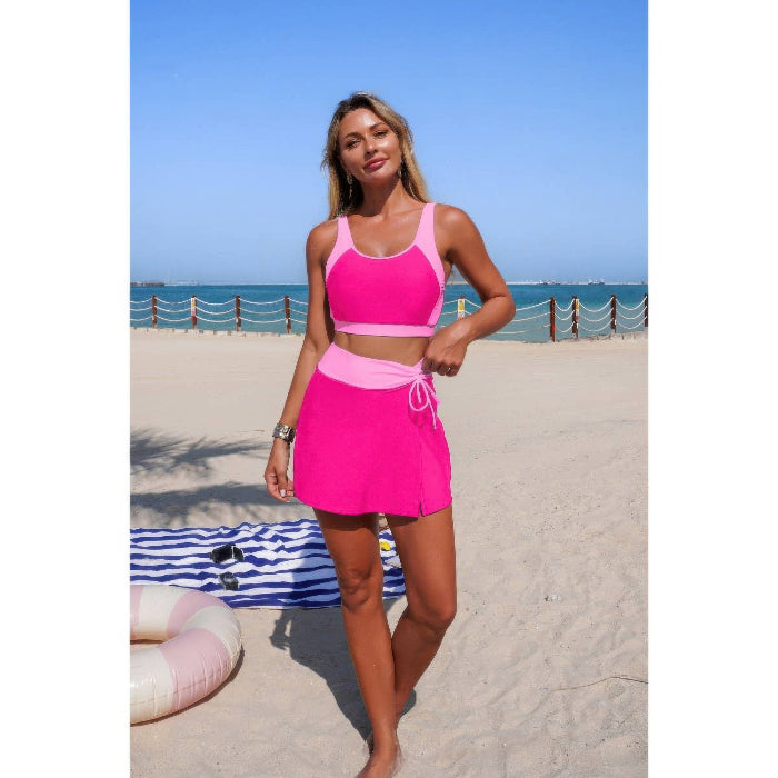 A woman modeling a bright pink two-piece swimsuit with a matching high-waisted tie-waist skirt on a sandy beach with the ocean and blue sky in the background.