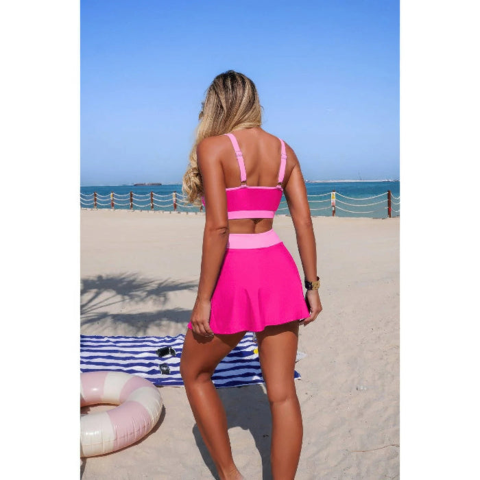 A woman stands on a sandy beach facing away from the camera, wearing a bright pink two-piece swimsuit with a strappy crop top and a flared mini skirt. The background shows the blue ocean, sky, and a striped beach towel.