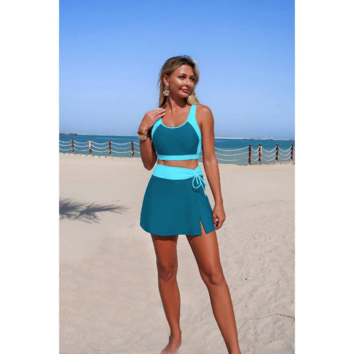 Woman in teal two-piece swimsuit standing on a sandy beach with ocean view.
