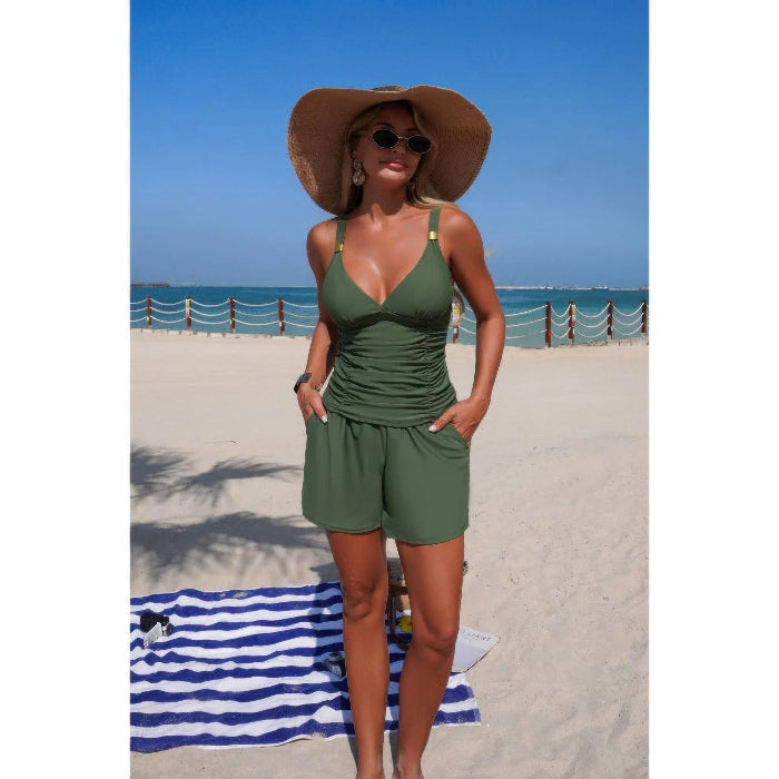 A woman stands on a sandy beach wearing an olive green two-piece swimsuit with a wide-brimmed straw hat and sunglasses, with the ocean and blue sky in the background.