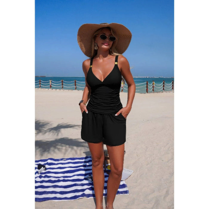 A woman stands on a sandy beach wearing a black one-piece swimsuit with shoulder straps, a large brown sun hat, and sunglasses. The background shows the blue ocean and sky.
