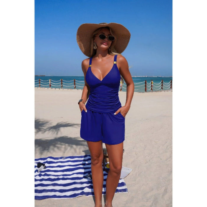 A woman stands on a sandy beach wearing a royal blue one-piece swimsuit with a wide-brimmed straw hat and sunglasses. The background shows the blue ocean and sky.