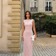 A woman stands outdoors on a cobblestone street wearing a sleeveless, fitted midi dress in a soft blush pink lace or textured fabric. 
