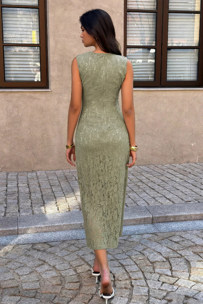 A woman wearing a sleeveless, knee-length sage green lace dress, photographed from behind in an outdoor setting with a stone-paved ground and a beige stucco wall background.