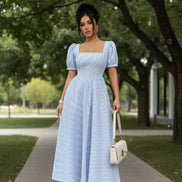 A woman models a light blue and white vertically striped midi dress with puff sleeves and a square neckline. She is holding a small cream-colored handbag an