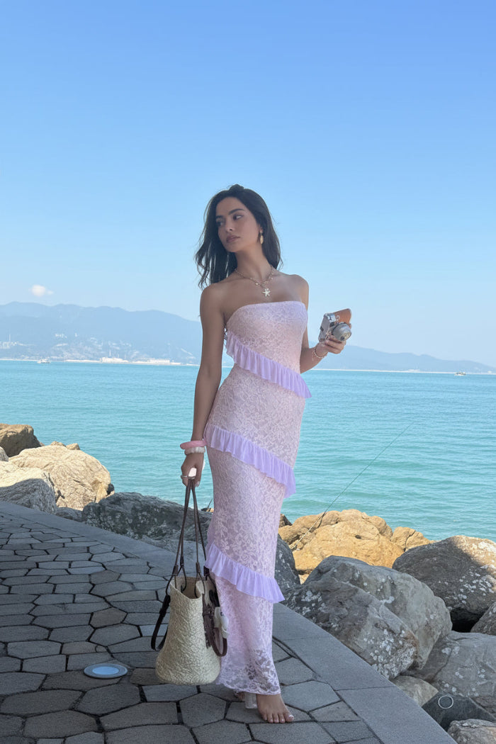 Woman in a pink lace dress standing by a coastal area with blue water and rocks.