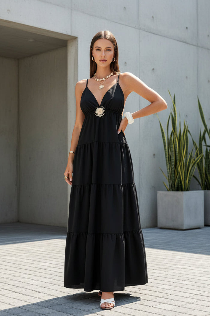 A woman wearing a black spaghetti-strap maxi dress with a tiered skirt and a silver sunburst embellishment at the bust, seated outdoors on a stone patio.