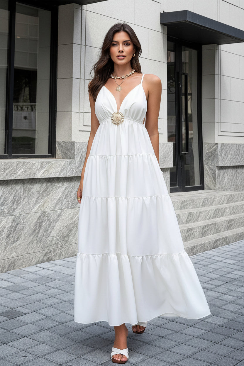 A woman wearing a white tiered maxi dress with thin straps, a V-neckline, and a decorative gold sunburst detail at the waist, standing outdoors on a paved walkway in front of a stone building.