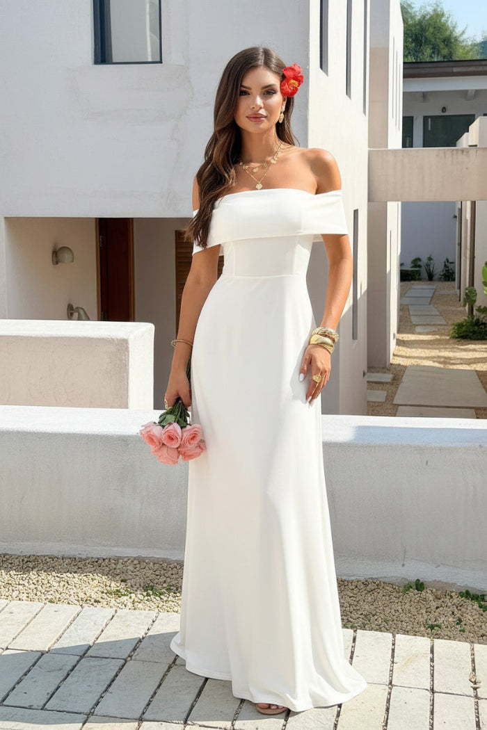 A woman wearing an off-the-shoulder white maxi dress with a fitted bodice and flowing skirt, leaning against a concrete wall outdoors while holding a bouquet of pink roses.