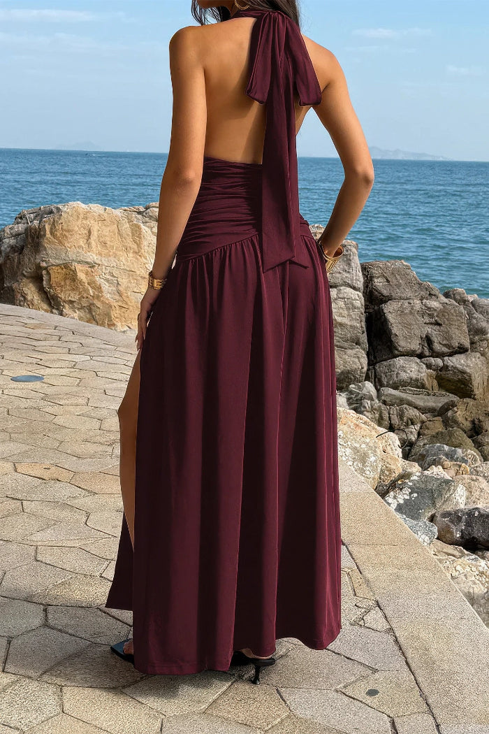 Woman in a burgundy halter neck dress standing by a rocky coastline.