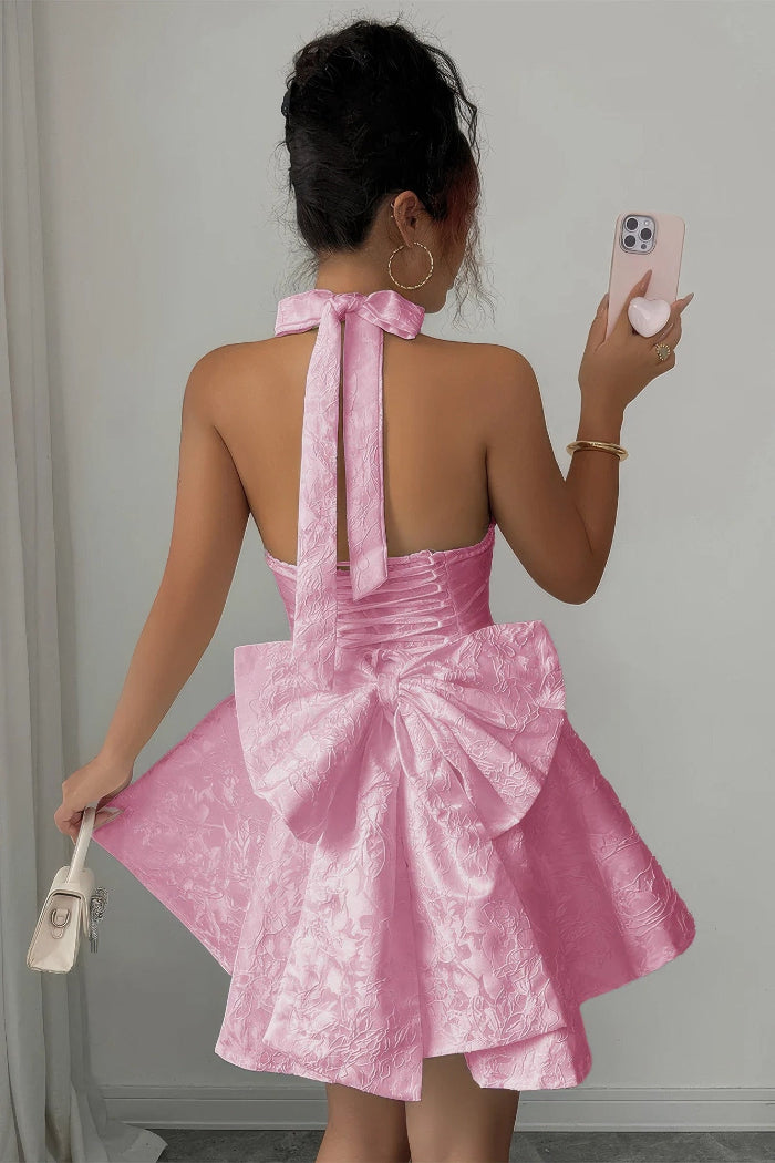 A woman photographed from behind wearing a short pink dress with a large bow at the back and a matching bow tie at the neck. She is holding a phone and a small handbag in an indoor setting.