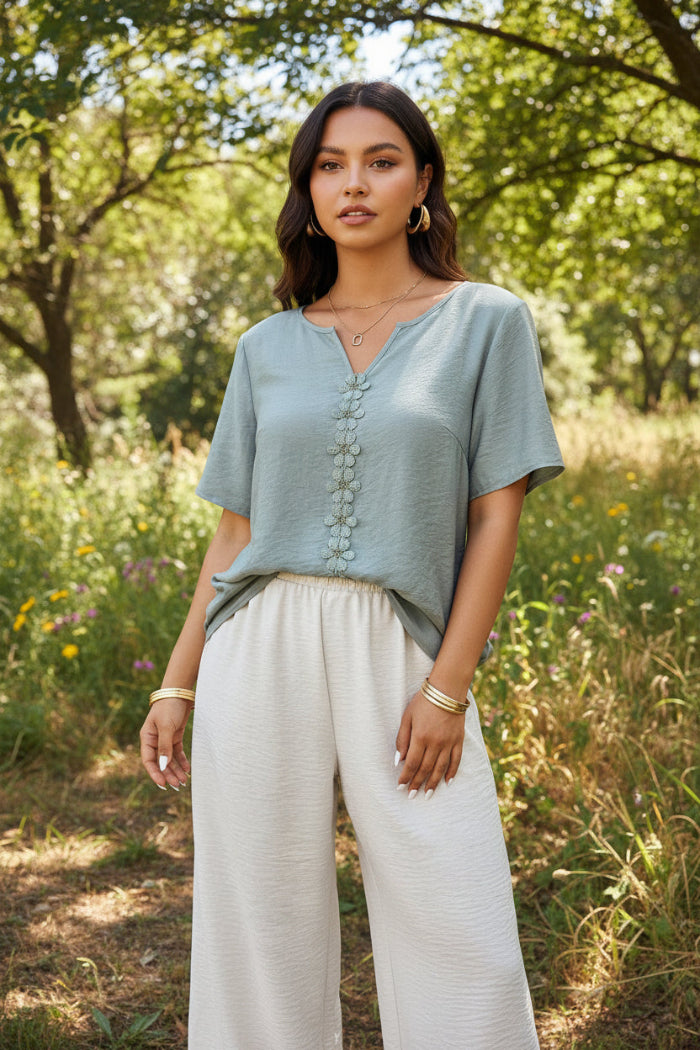 Woman wearing a light gray top with decorative buttons and beige pants indoors.