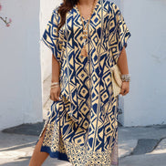 A woman wearing a loose, knee-length kaftan dress with a navy blue and beige geometric pattern, styled with gold jewelry and beige sandals, standing in front of a white wall.