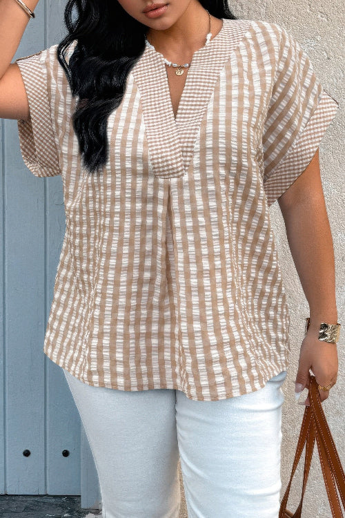 Woman standing outdoors in a loose khaki-and-white textured plaid blouse with a split V-neck and striped sleeve trim, paired with white pants and a brown handbag
