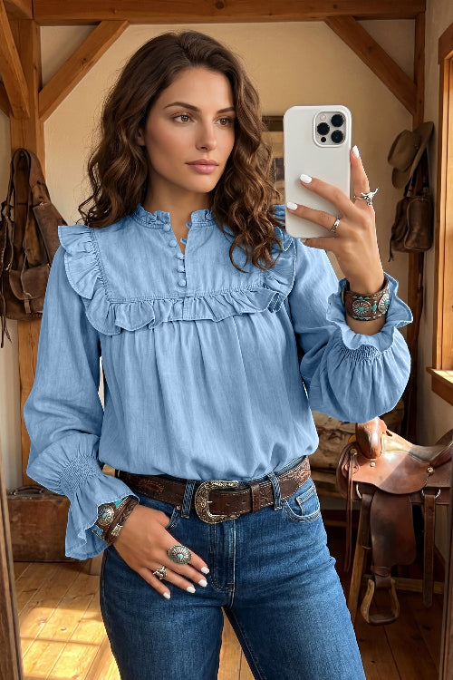 A woman takes a mirror selfie wearing a light blue long-sleeve blouse with ruffle details, paired with dark blue jeans and a brown belt, in a rustic indoor setting with wooden beams and saddles.