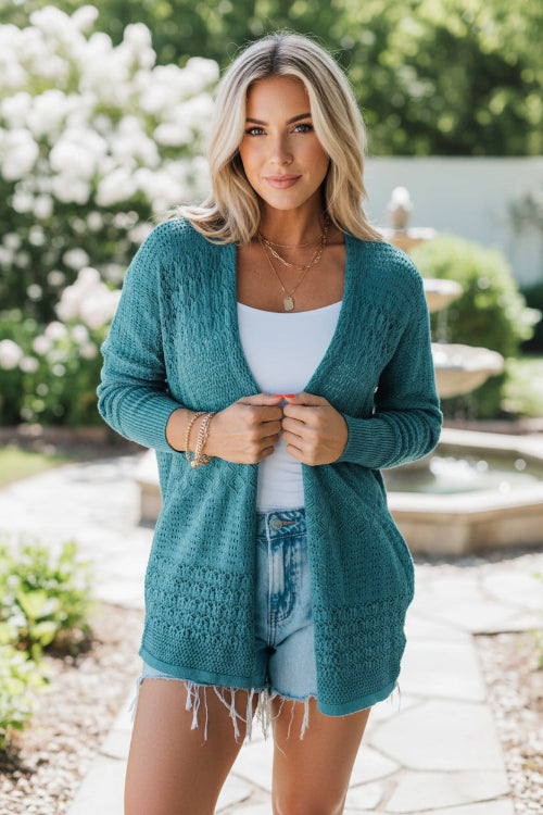 Woman wearing a teal cardigan over a white top and denim shorts against a light gray background