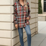 A woman wearing a red, brown, and blue plaid hooded button-up shirt leans against a beige stone wall, holding a coffee cup.