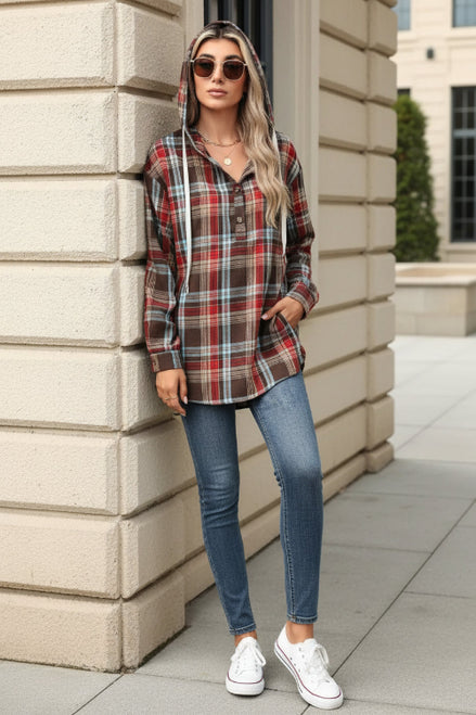 A woman wearing a red, brown, and blue plaid hooded button-up shirt leans against a beige stone wall, holding a coffee cup.