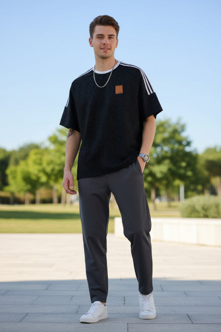 Man wearing a black t-shirt with white stripes and gray pants standing outdoors on a sunny day.