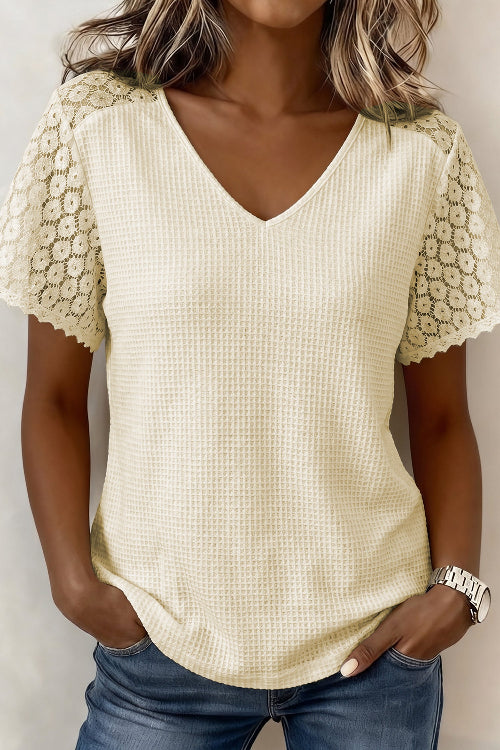 A woman wearing a cream-colored waffle-knit V-neck top with floral lace short sleeves, paired with blue jeans.
