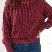 A woman wearing a ribbed knit long-sleeve sweater in a deep burgundy red, paired with blue jeans and a delicate silver necklace.