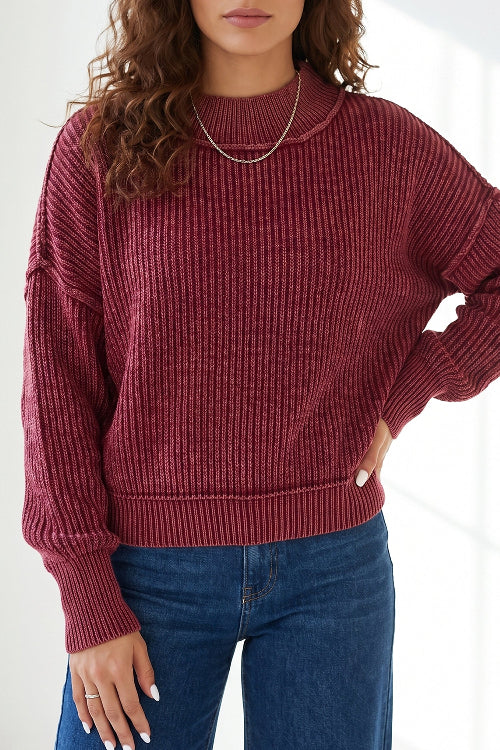 A woman wearing a ribbed knit long-sleeve sweater in a deep burgundy red, paired with blue jeans and a delicate silver necklace.