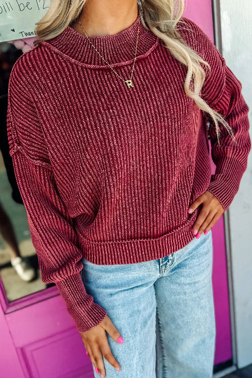 A woman wearing a ribbed knit crewneck sweater in a deep burgundy red, paired with light blue jeans and a gold necklace, 
