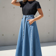 Person wearing a black t-shirt and blue denim skirt against a white wall.