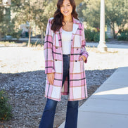 A woman wearing a pink, white, and black plaid double-breasted coat over a white tank top and dark blue jeans, 