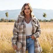 Person wearing a plaid jacket over a white top and blue jeans, holding a beige handbag against a gray background.