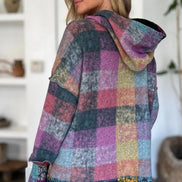 Woman wearing a multicolored checkered hoodie in a room with white walls and a plant.