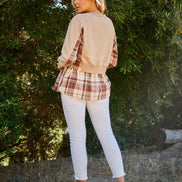 Woman wearing a beige sweater over a plaid shirt with white pants, standing in a natural setting.
