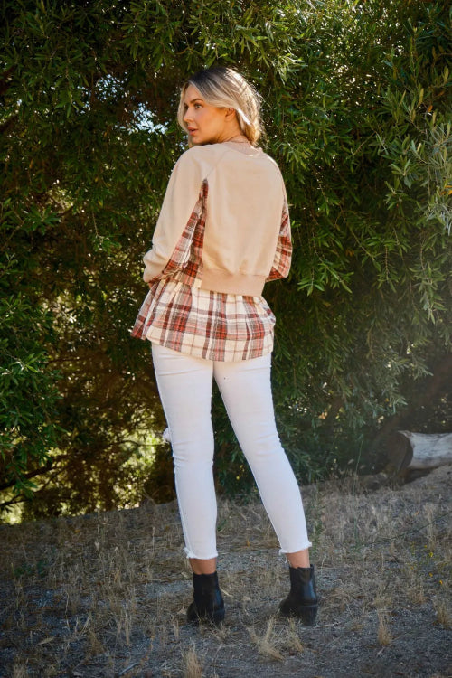 Woman wearing a beige sweater over a plaid shirt with white pants, standing in a natural setting.