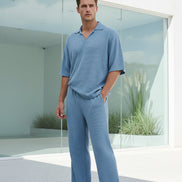 Man wearing a light blue outfit standing on a wooden deck with a white wall and greenery in the background.