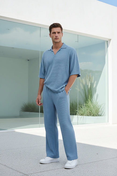 Man wearing a light blue outfit standing on a wooden deck with a white wall and greenery in the background.