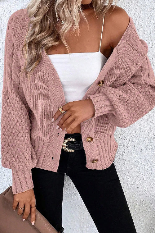 A woman wearing a dusty pink chunky knit cardigan with gold buttons over a white camisole and black jeans, standing against a white textured wall.