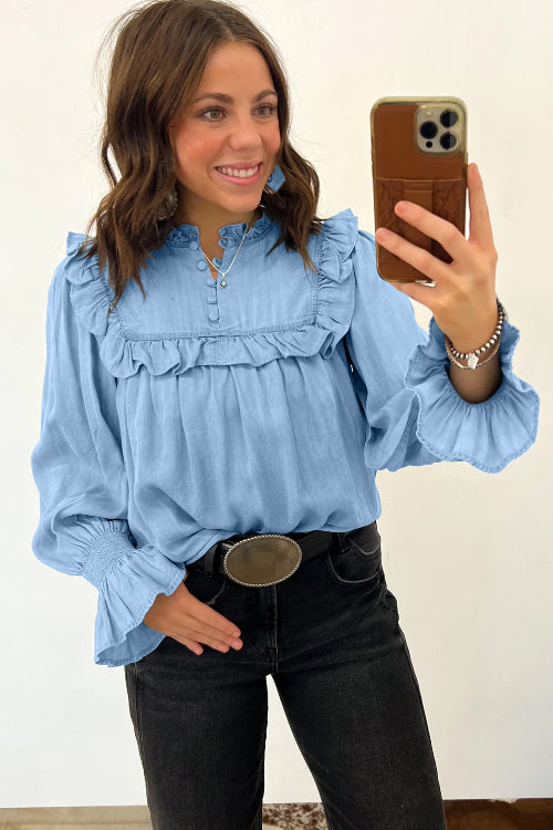 A woman takes a mirror selfie wearing a light blue long-sleeve blouse with ruffle details, paired with black jeans and a silver belt.