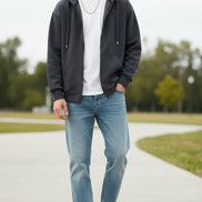 Man wearing a dark hoodie, white t-shirt, light blue jeans, and white sneakers standing outdoors.