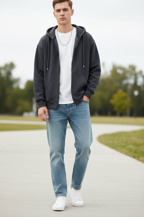 Man wearing a dark hoodie, white t-shirt, light blue jeans, and white sneakers standing outdoors.