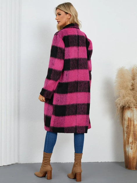 Woman wearing a pink and black checkered coat with blue pants and brown boots, standing against a white wall.