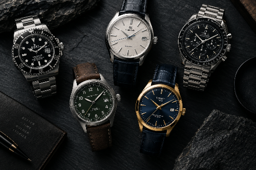 Six luxury wristwatches arranged on a dark textured surface with rocks and a notebook. The watches include stainless steel, silver, green, gold, and blue 
